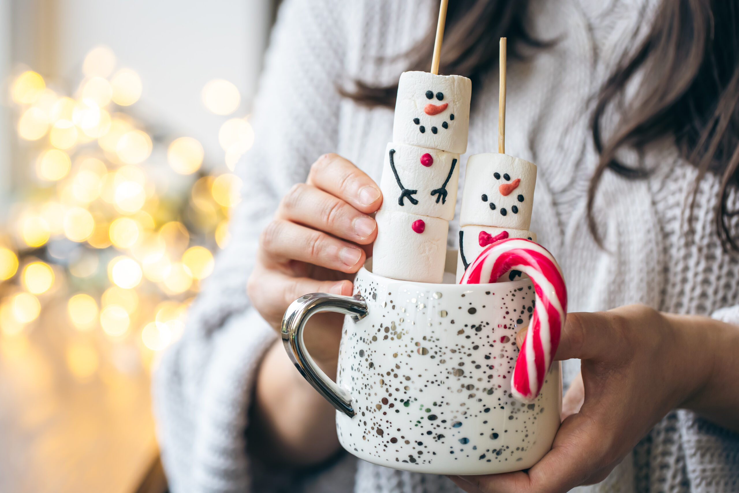 Cup of hot drink with marshmallow snowmen and candy in female hands. Hot cocoa with marshmallow snowmen and candy cane, festive Christmas treat that can contribute to holiday tooth staining