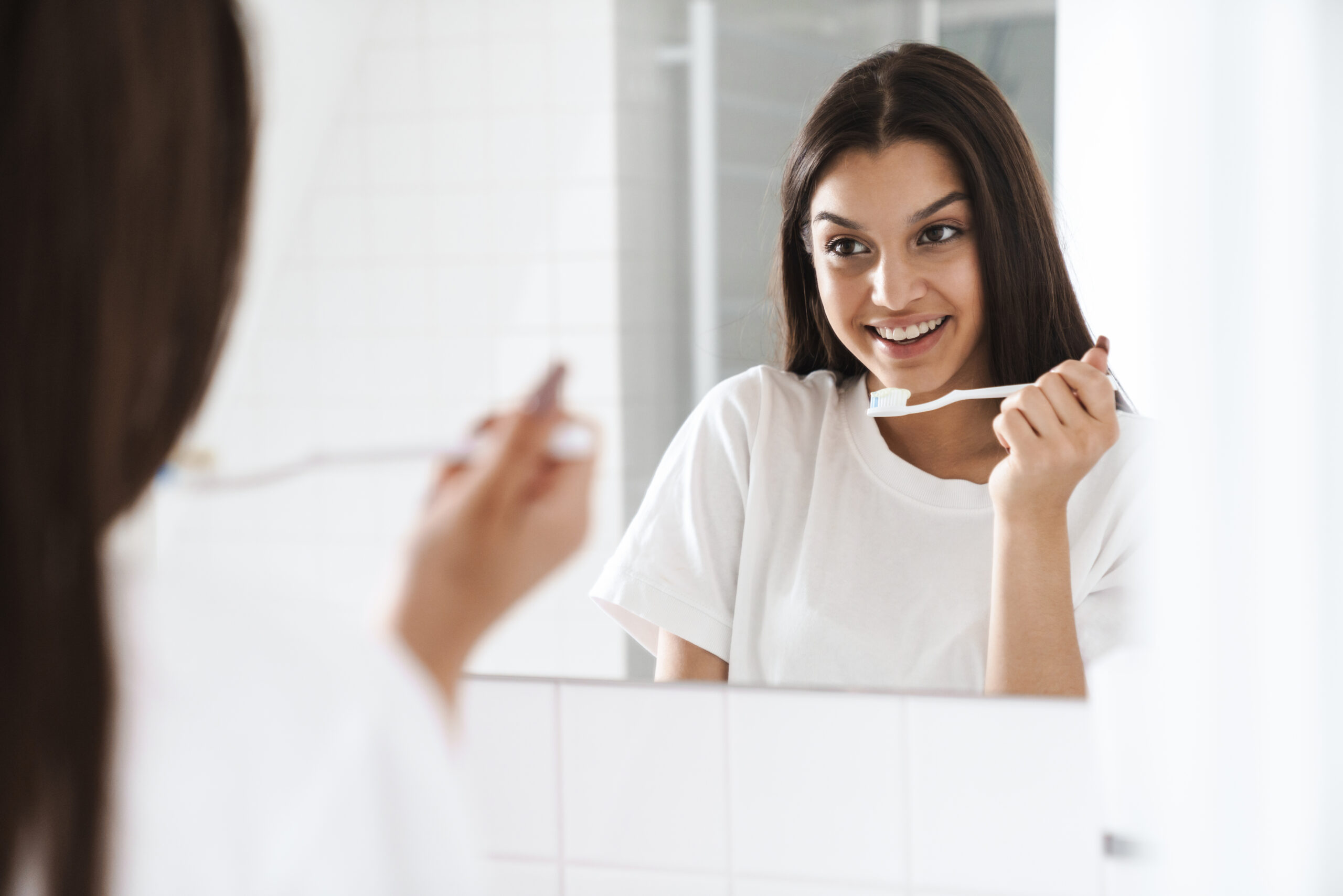 Photo of woman smiling and cleaning teethes while looking at mirror Woman brushing her teeth in front of a mirror, demonstrating a healthy daily oral care routine at home